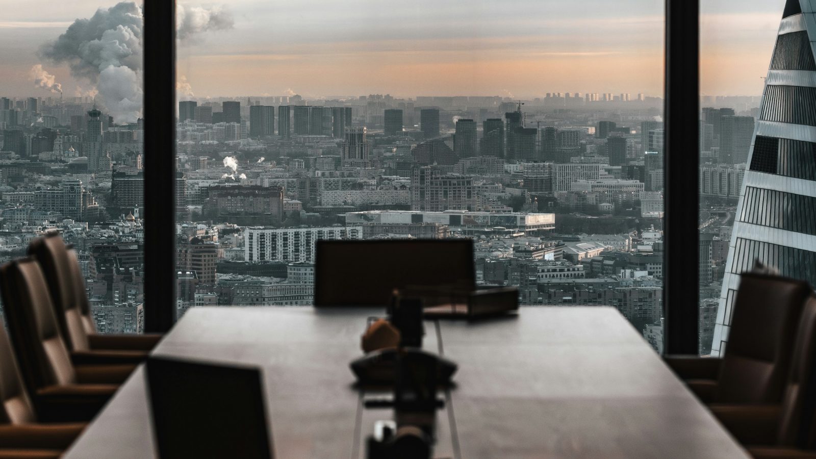 An empty boardroom table with chairs, facing a city skyline at dusk